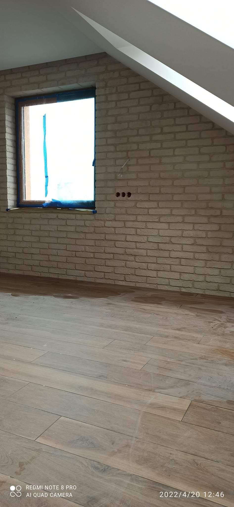 Pokój na poddaszu z wykończoną ścianą z cegły dekoracyjnej w odcieniach beżu, ułożonymi panelami podłogowymi i oknem zabezpieczonym niebieską folią malarską.