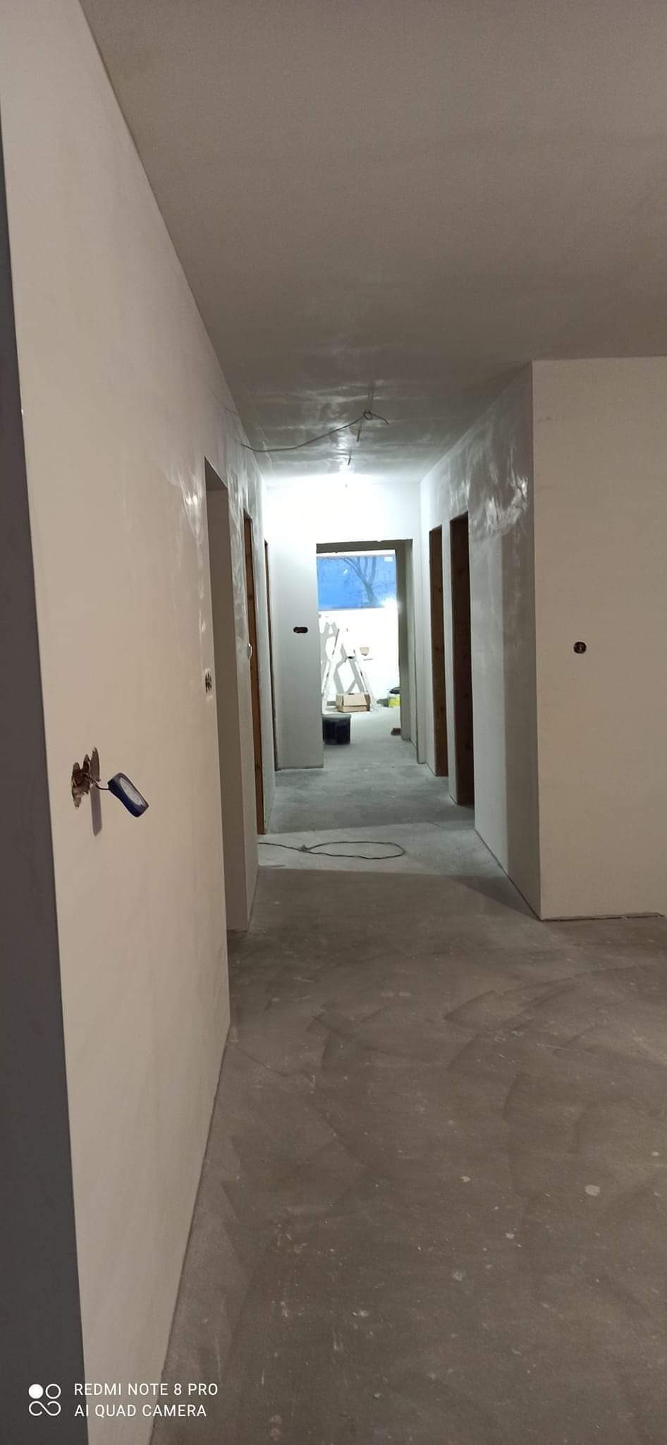 Korytarz w trakcie prac wykończeniowych, ściany pokryte świeżo nałożoną gładzią szpachlową, widoczne ślady po szpachli, przygotowane pod malowanie, w tle drabina i otwarte drzwi.