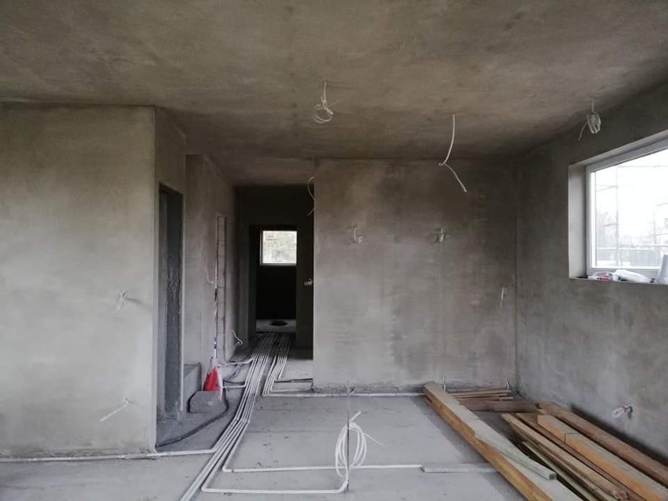 Surowe ściany w trakcie prac wykończeniowych, widoczne kable elektryczne zwisające z sufitu oraz ułożone na podłodze rury instalacyjne i deski.