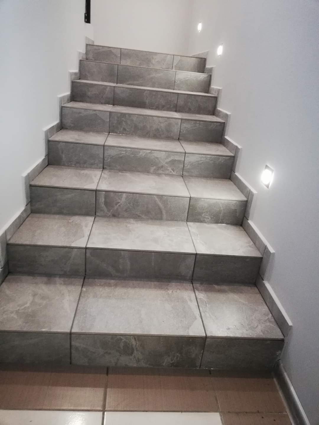 Schody wewnętrzne wyłożone szarymi płytkami ceramicznymi z subtelnym wzorem marmurkowym, oświetlone punktowymi lampami ściennymi, widok z dołu.