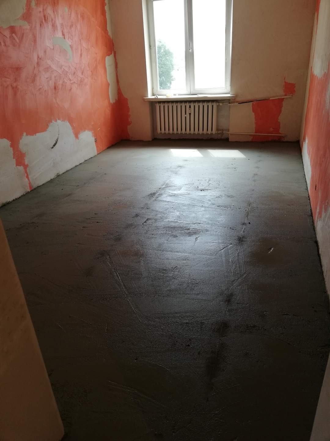 Świeżo wylana, mokra wylewka samopoziomująca na podłodze w pokoju podczas prac przygotowawczych do dalszych prac wykończeniowych, ściany częściowo pokryte pomarańczową farbą i gładzią, widoczne...