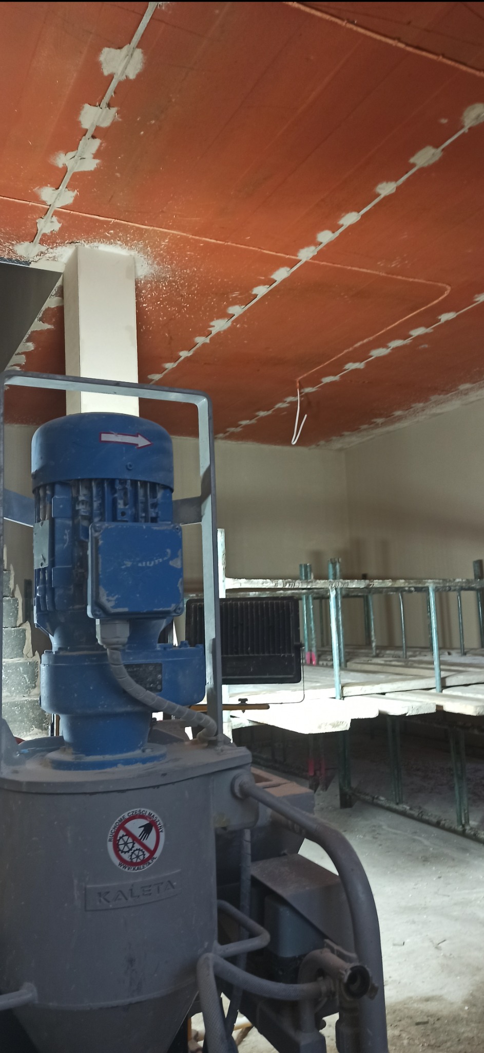 Widok na maszynę do tynkowania Kaleta, z surowym, pomarańczowym sufitem w tle, gdzie widoczne są ślady szpachlowania połączeń płyt.