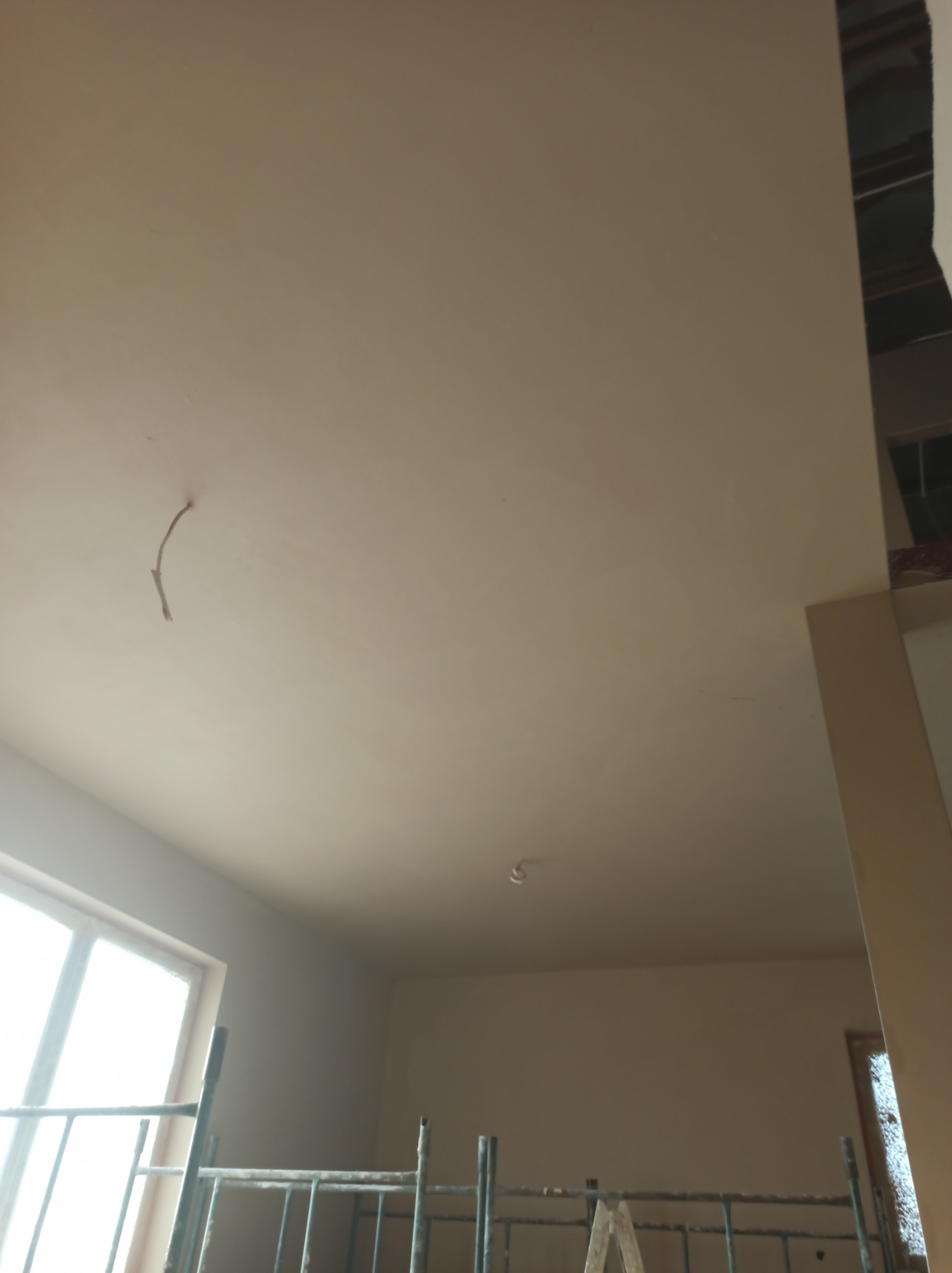 Wnętrze w trakcie prac wykończeniowych, widoczny sufit po nałożeniu gładzi, z wystającym przewodem elektrycznym, oraz fragment ściany. W tle widoczne okno i rusztowanie.