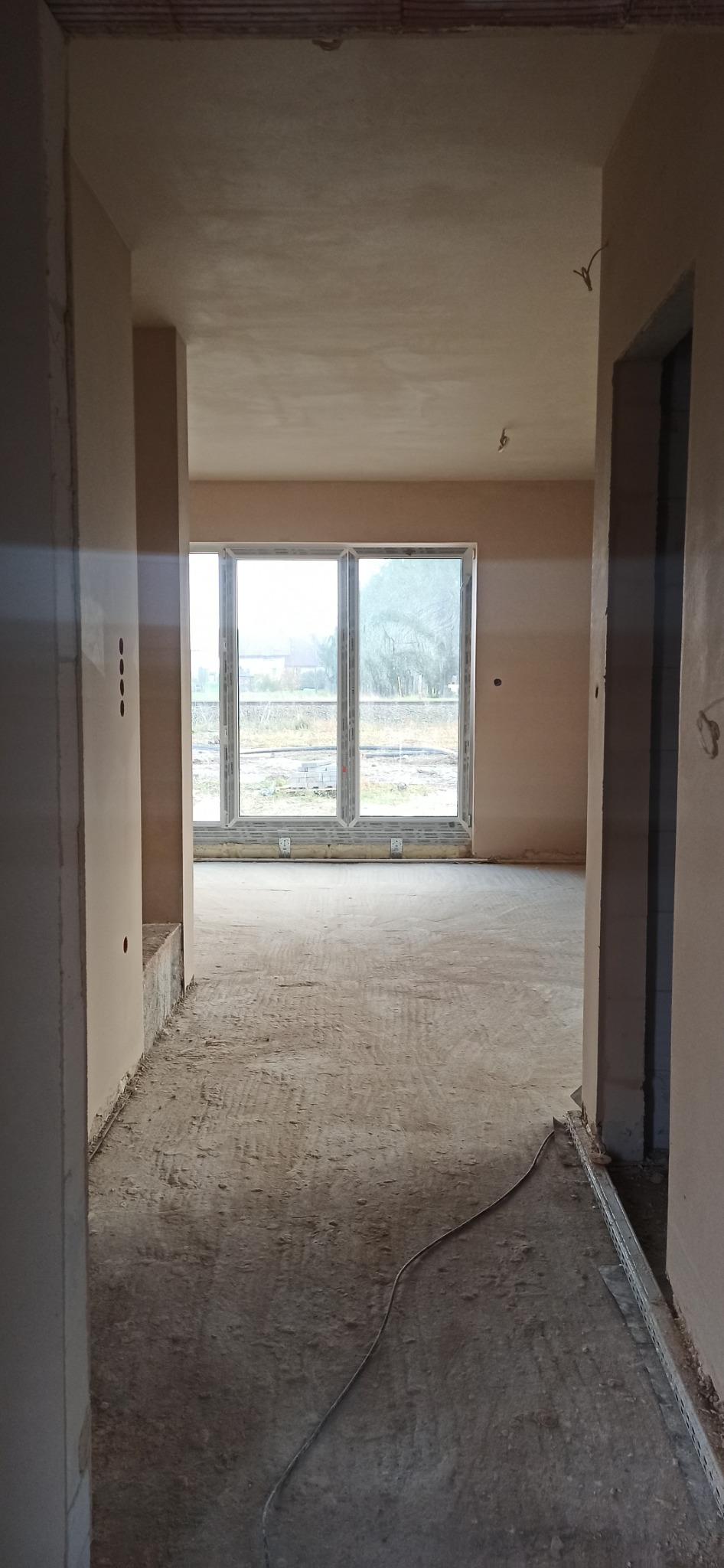 Wnętrze w trakcie prac wykończeniowych, ściany i sufit pokryte świeżym tynkiem, widoczne okno z widokiem na zewnątrz, kabel na podłodze i profile podłogowe.