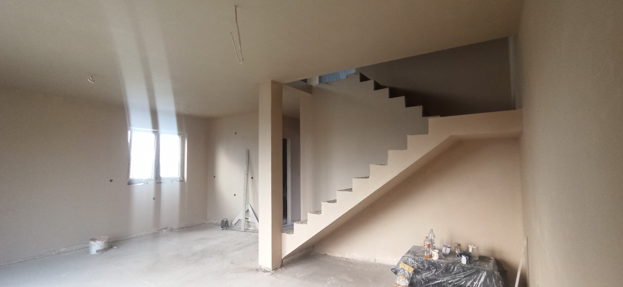 Wnętrze domu w trakcie prac wykończeniowych: świeżo położone tynki, widoczne schody, okno z mocnym światłem dziennym, wiadro i narzędzia malarskie zabezpieczone folią.