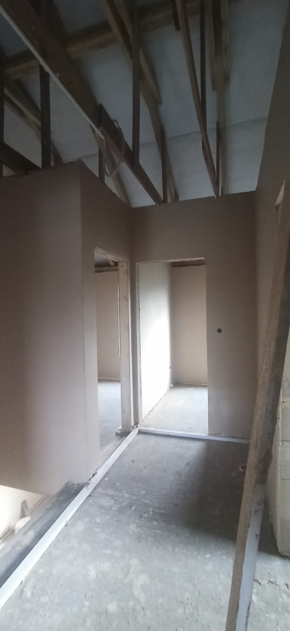 Surowe wnętrze poddasza z nałożonymi tynkami na ścianach, widoczne drewniane elementy konstrukcji dachu i przygotowane otwory drzwiowe.