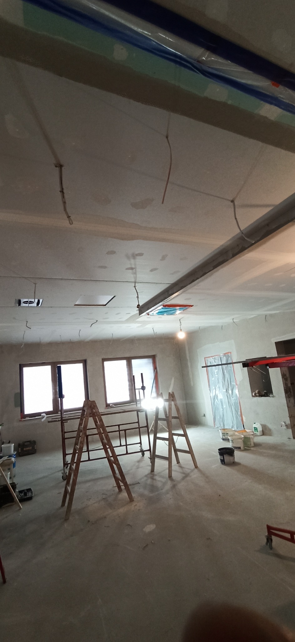 Wnętrze w trakcie remontu: sufit i ściany po szpachlowaniu, widoczne drabiny, rusztowanie, kable zwisające z sufitu, okna i zabezpieczone folią drzwi.
