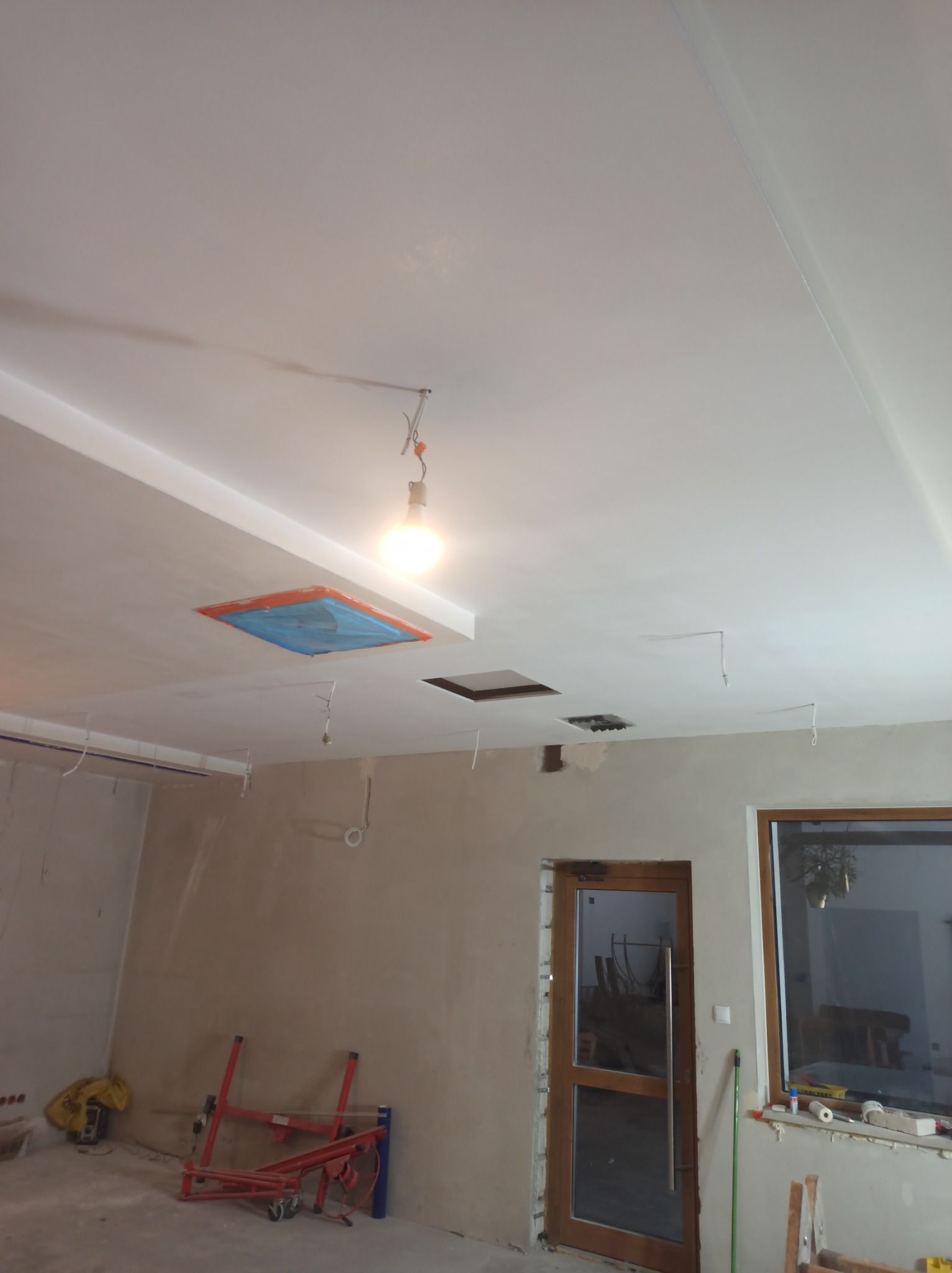 Wnętrze w trakcie prac wykończeniowych: surowa ściana, przygotowany otwór w suficie zabezpieczony folią, widoczne przewody elektryczne, drabina malarska i lampa wisząca na kablu.