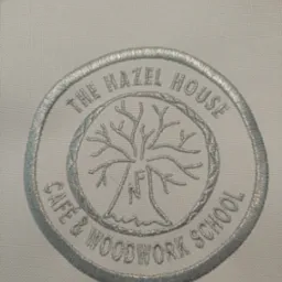 Jasnoszary, okrągły emblemat z haftowanym drzewem i napisem 'The Hazel House Cafe & Woodwork School' na białym materiale.