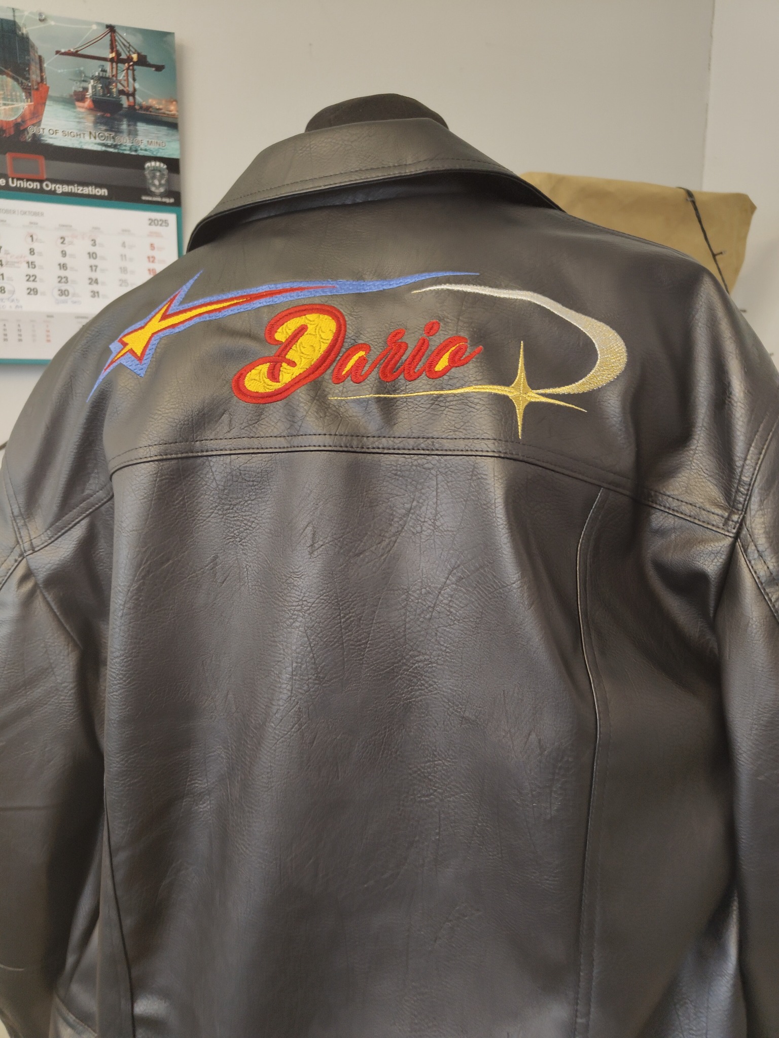 Czarna kurtka z ekoskóry z haftowanym napisem 'Dario', zdobiona motywem gwiazdy i błyskawicy w kolorach czerwonym, żółtym, niebieskim i srebrnym. Widok z tyłu.