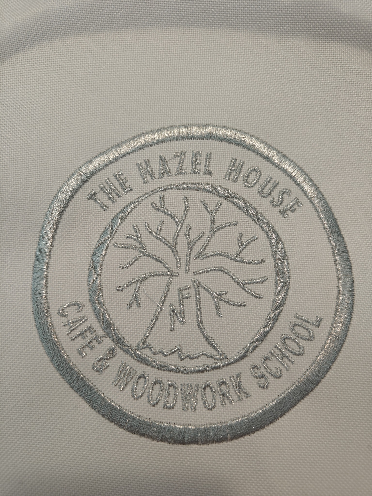 Jasnoszary, okrągły emblemat z haftowanym drzewem i napisem 'The Hazel House Cafe & Woodwork School' na białym materiale.