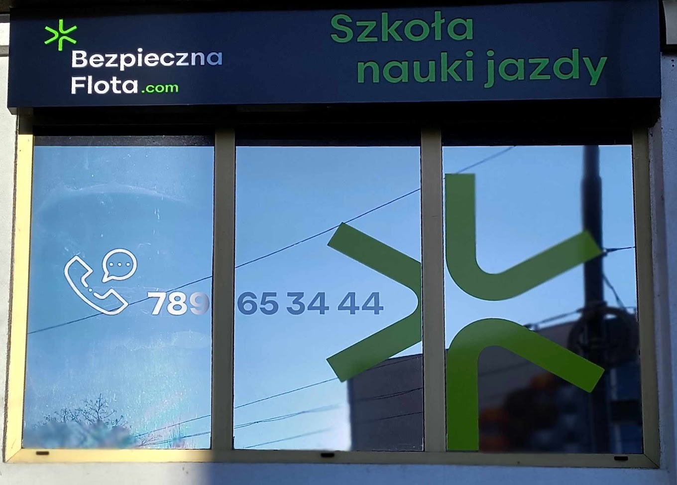 Fasada budynku z szyldem 'Szkoła nauki jazdy' i logo 'Bezpieczna Flota.com', numer telefonu naklejony na oknie.