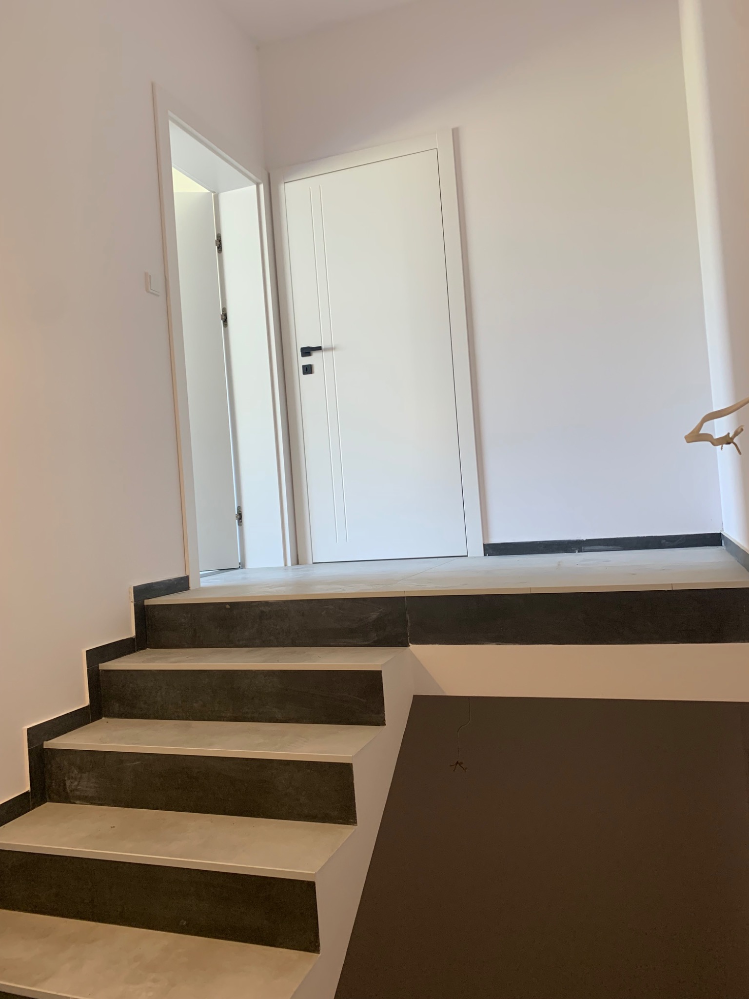 Nowoczesne schody wykończone szarym gresem, białe ściany i dwie białe drzwi z czarnymi klamkami w nowym budynku.