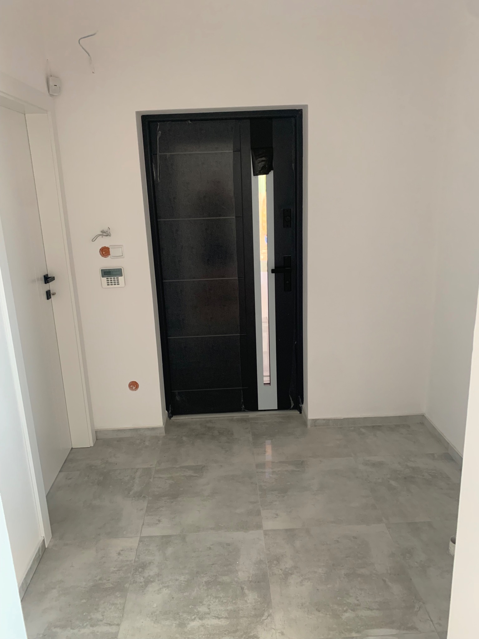 Nowoczesne wejście do mieszkania z czarnymi drzwiami i szarą podłogą z płytek imitujących beton, widoczne niedokończone podłączenia instalacji alarmowej.