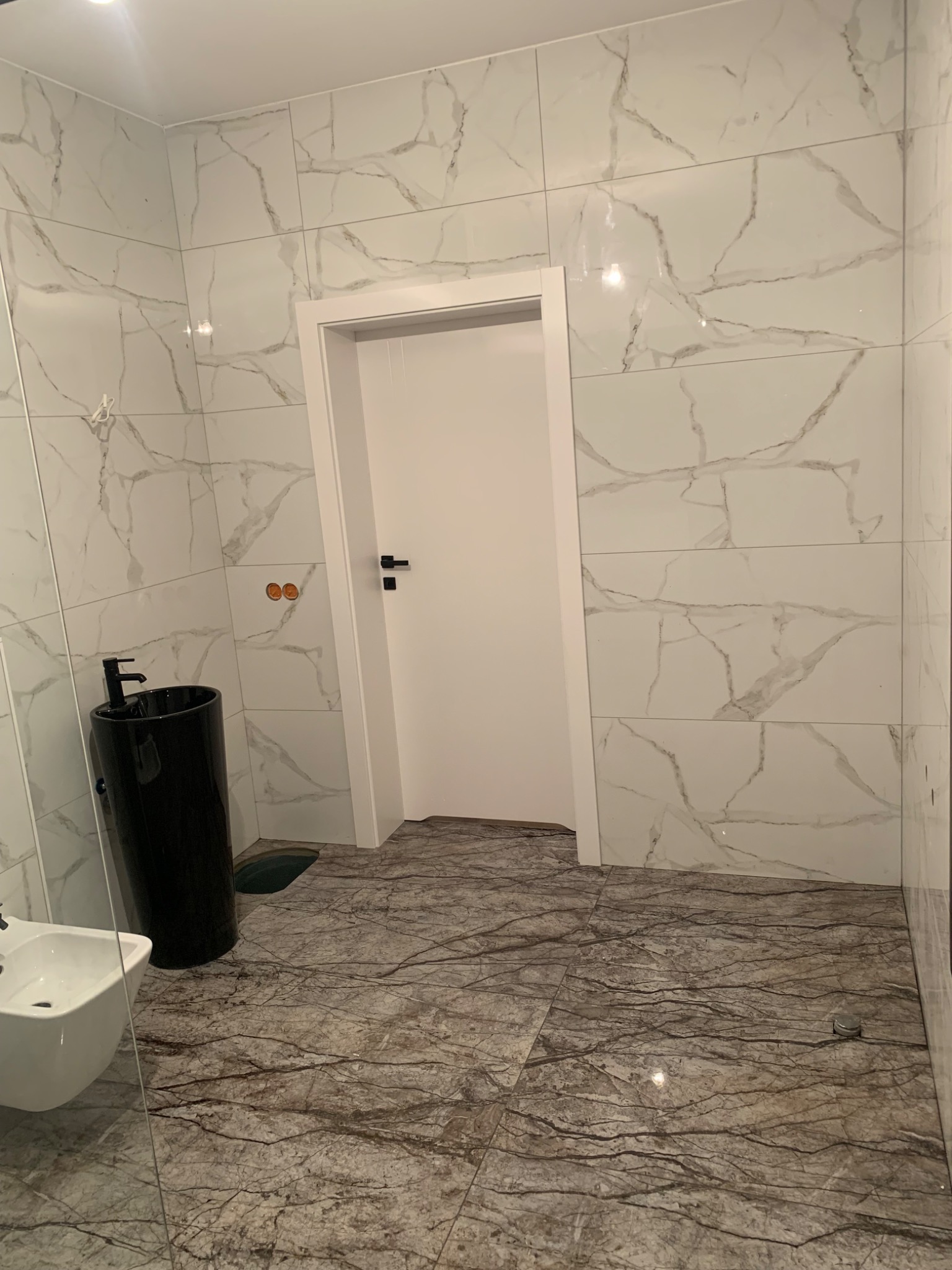 Wykończona łazienka z białymi płytkami imitującymi marmur na ścianach, ciemnobrązowymi płytkami podłogowymi z wzorem żyłkowania, czarną umywalką na postumencie i białą miską bidetową.