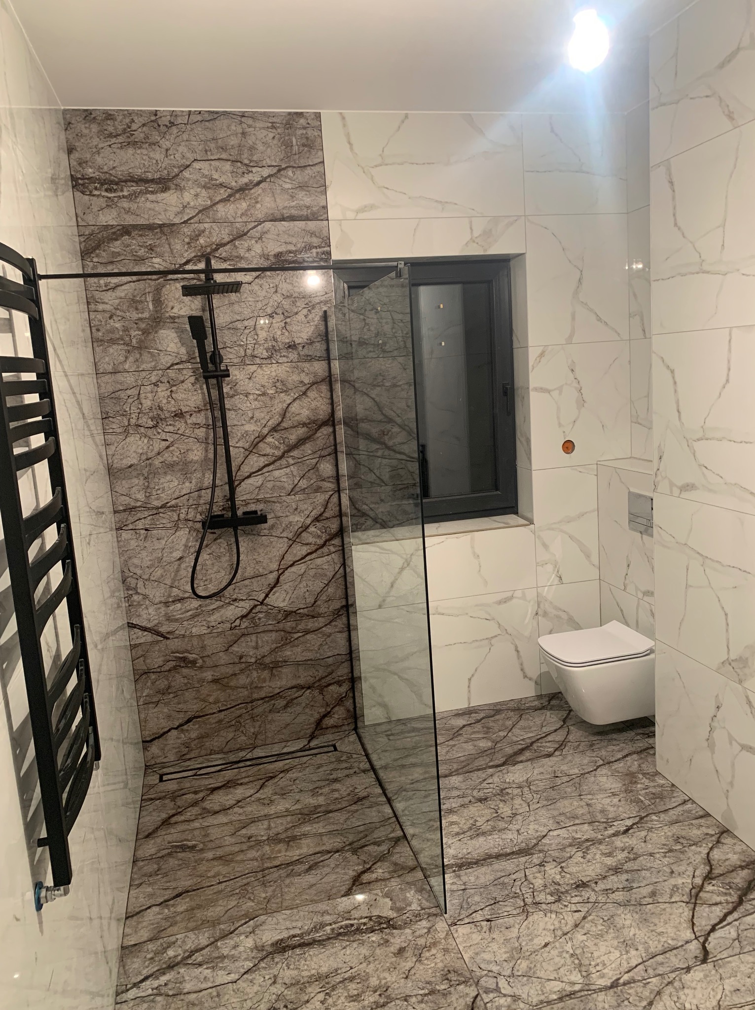 Nowoczesna łazienka z kabiną prysznicową typu walk-in, czarną armaturą i grzejnikiem drabinkowym, marmurowe płytki na podłodze i ścianach, wisząca toaleta.