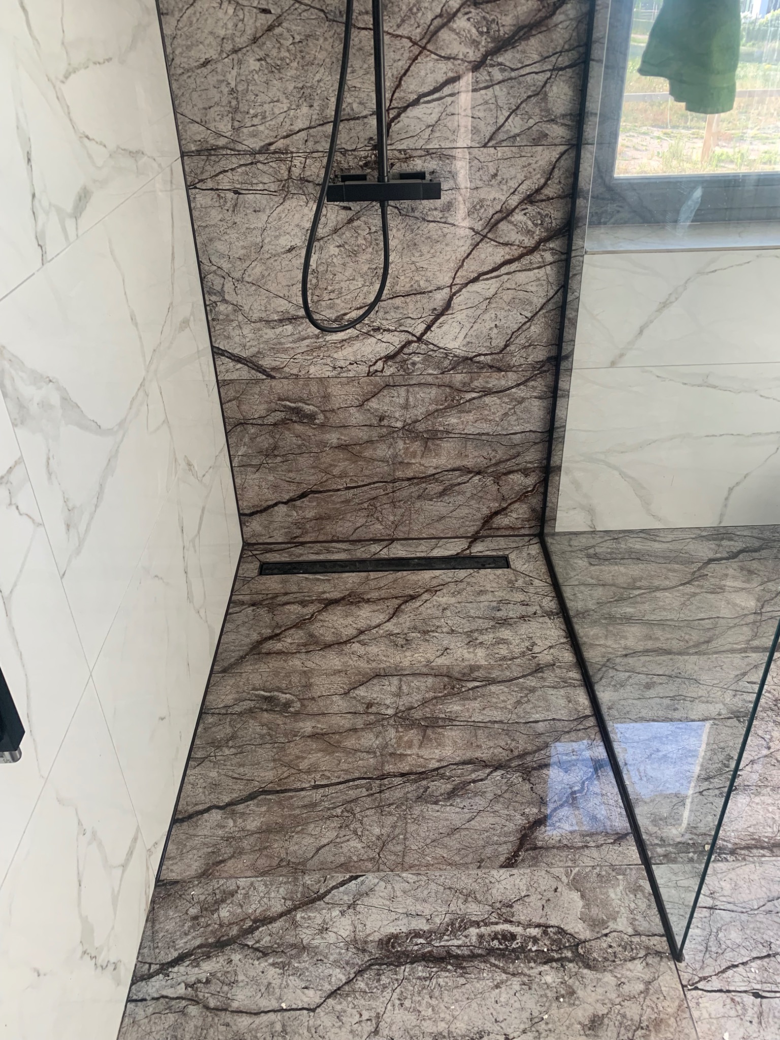 Nowoczesna kabina prysznicowa walk-in z marmurowymi płytkami w odcieniach szarości i brązu, kontrastująca z białą ścianą wyłożoną płytkami imitującymi marmur, czarna armatura prysznicowa i odpływ...