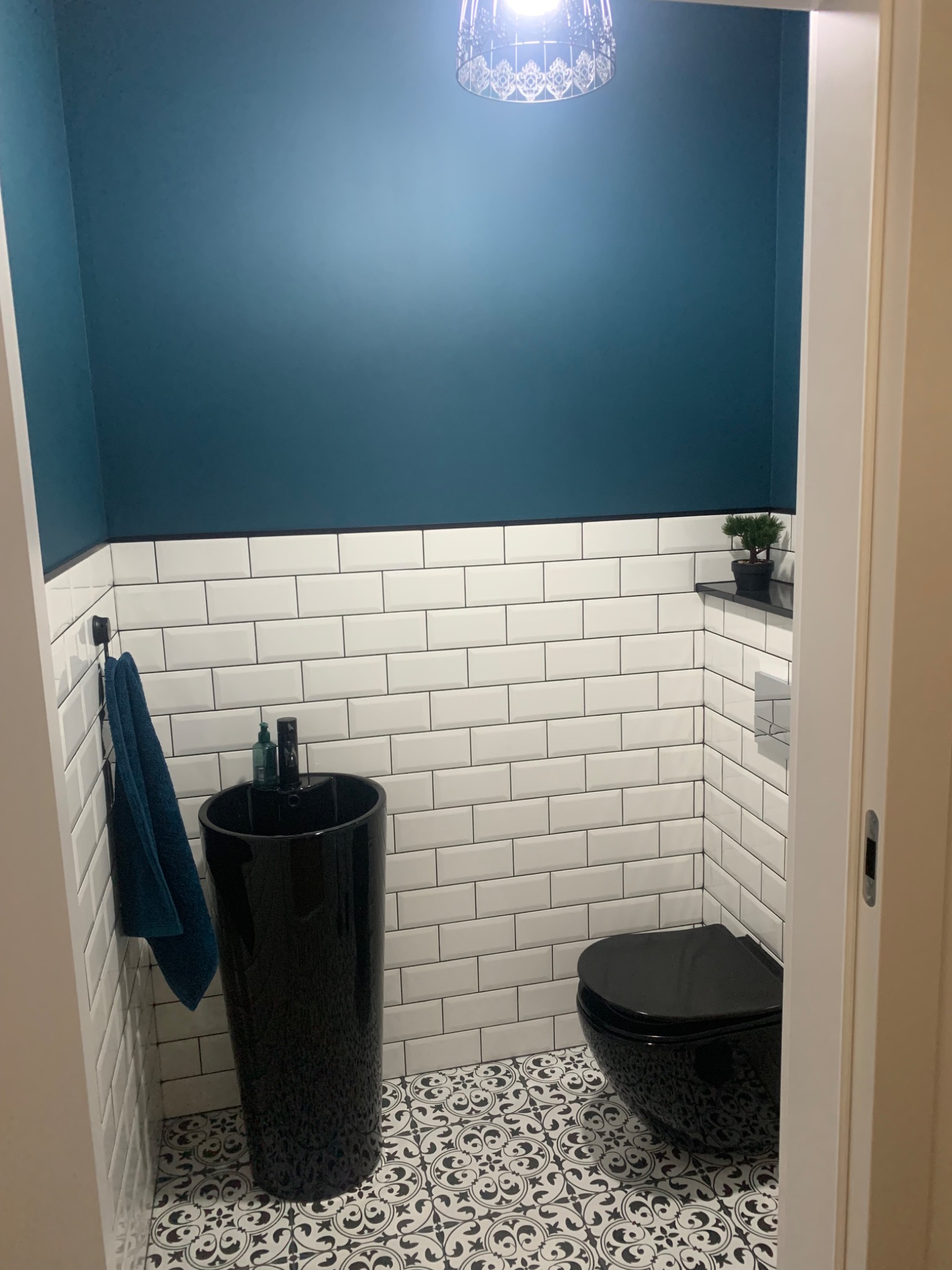 Mała toaleta z ciemnoniebieską ścianą, białymi płytkami typu metro z czarną fugą, czarną umywalką kolumnową, wiszącą toaletą w tym samym kolorze i wzorzystą podłogą w stylu retro.
