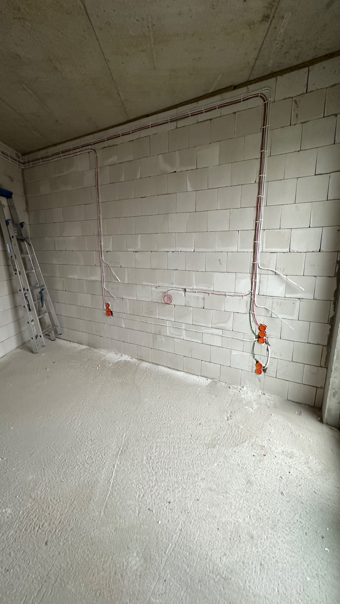 Surowy stan wnętrza z widoczną instalacją elektryczną: przewody w białych listwach, pomarańczowe puszki podtynkowe, drabina oparta o ścianę z betonu komórkowego.