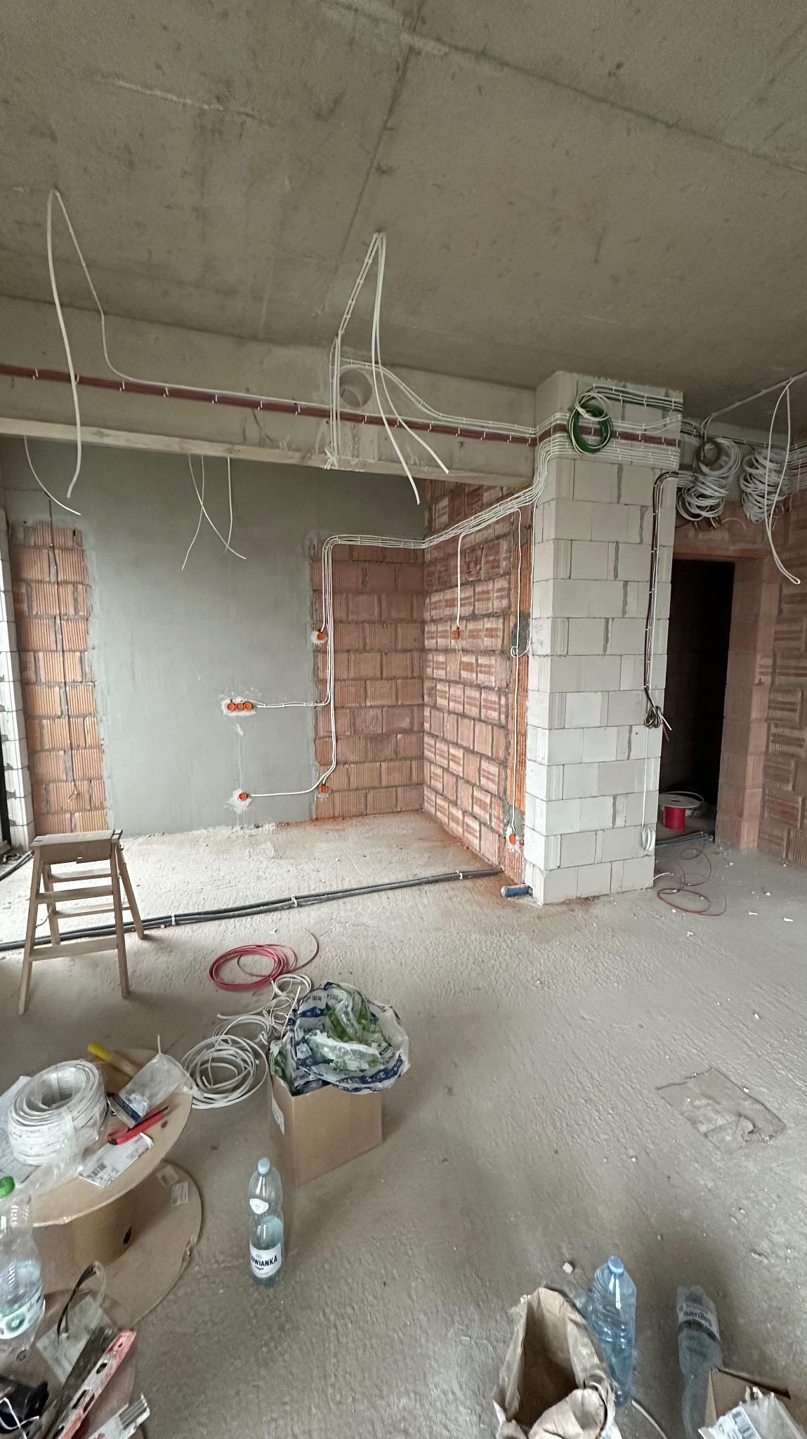 Wnętrze w trakcie remontu z widoczną instalacją elektryczną: kable prowadzone po ścianach z cegły i betonu, puszki elektryczne zamontowane, przewody zwisające z sufitu, widoczne materiały budowlane...