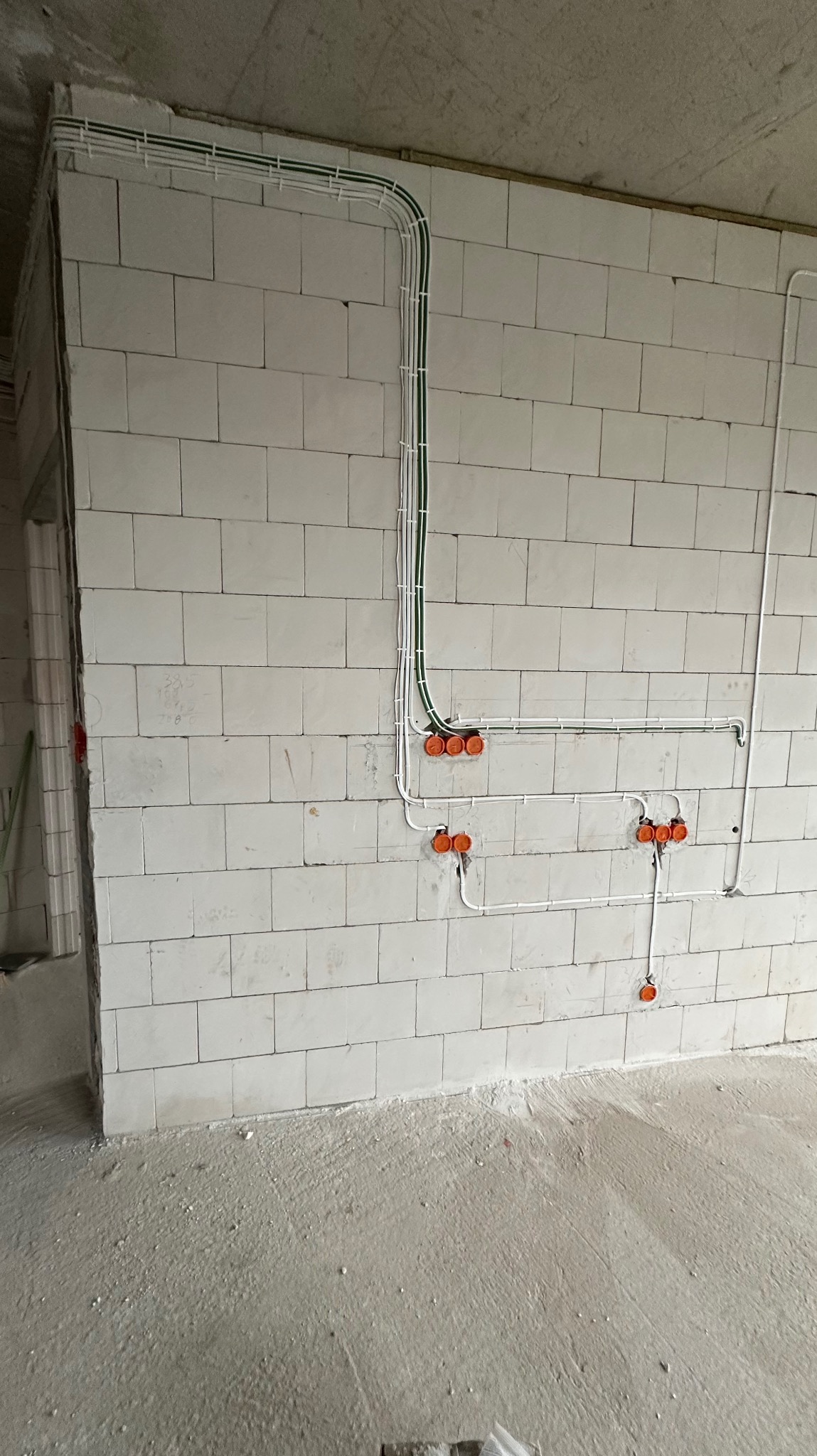 Surowe wnętrze w trakcie prac wykończeniowych: widoczne przewody elektryczne w białych rurkach i pomarańczowe puszki instalacyjne zamontowane na ścianie z białych bloczków.
