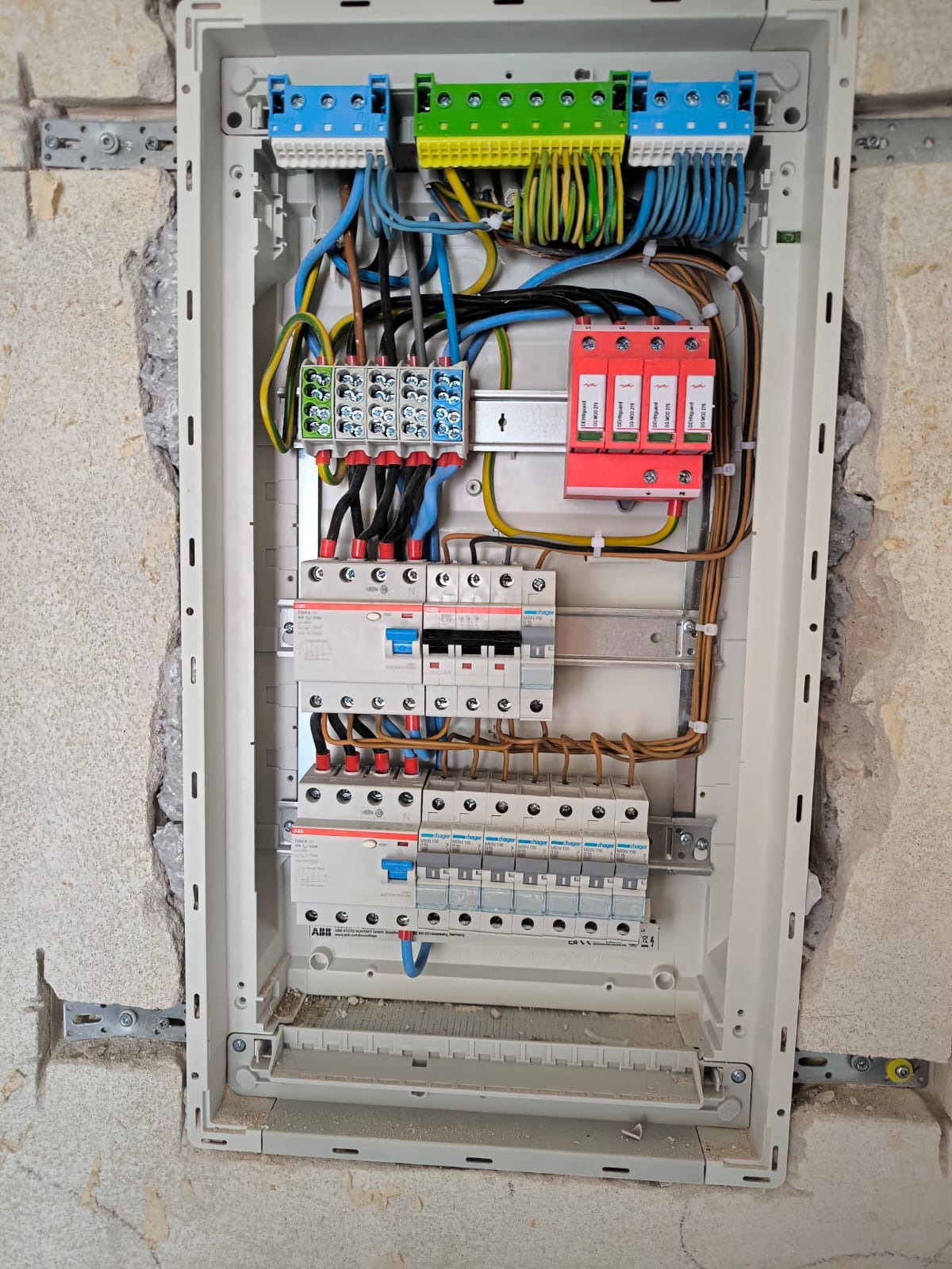 Otwarta skrzynka elektryczna z różnokolorowymi przewodami, bezpiecznikami i listwami zaciskowymi, zamontowana na surowej ścianie z widocznymi śladami po kuciu.