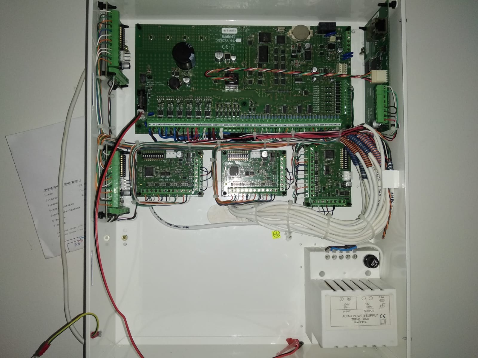 Otwarta skrzynka z centralą alarmową Satel Integra 64, widoczne moduły elektroniczne, liczne przewody, zasilacz oraz kartka z odręcznymi notatkami po lewej stronie.