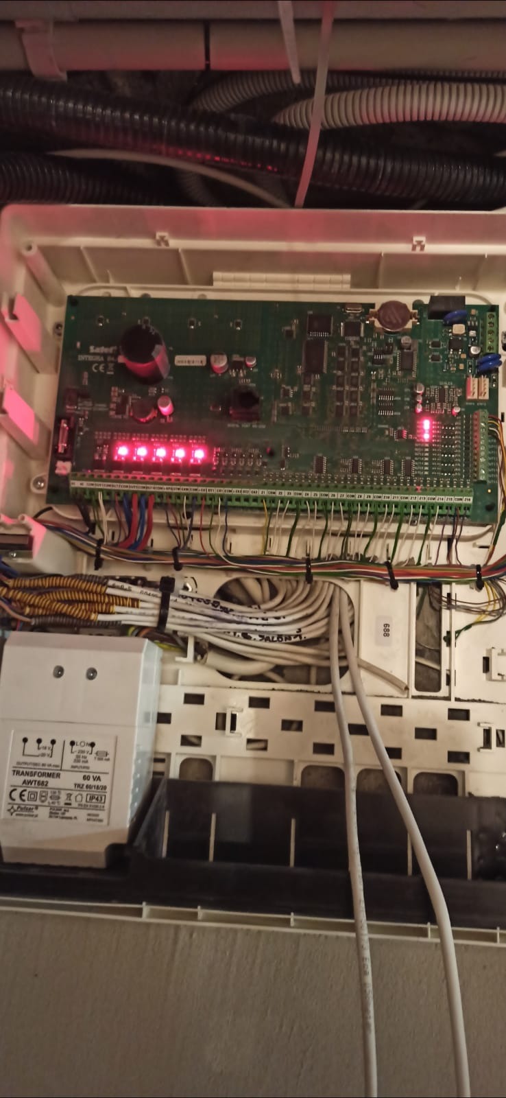 Otwarta obudowa centrali alarmowej Satel Integra 64 z widoczną płytą główną, licznymi przewodami podłączonymi do listew zaciskowych, transformatorem AWT682 i diodami LED sygnalizującymi stan systemu.