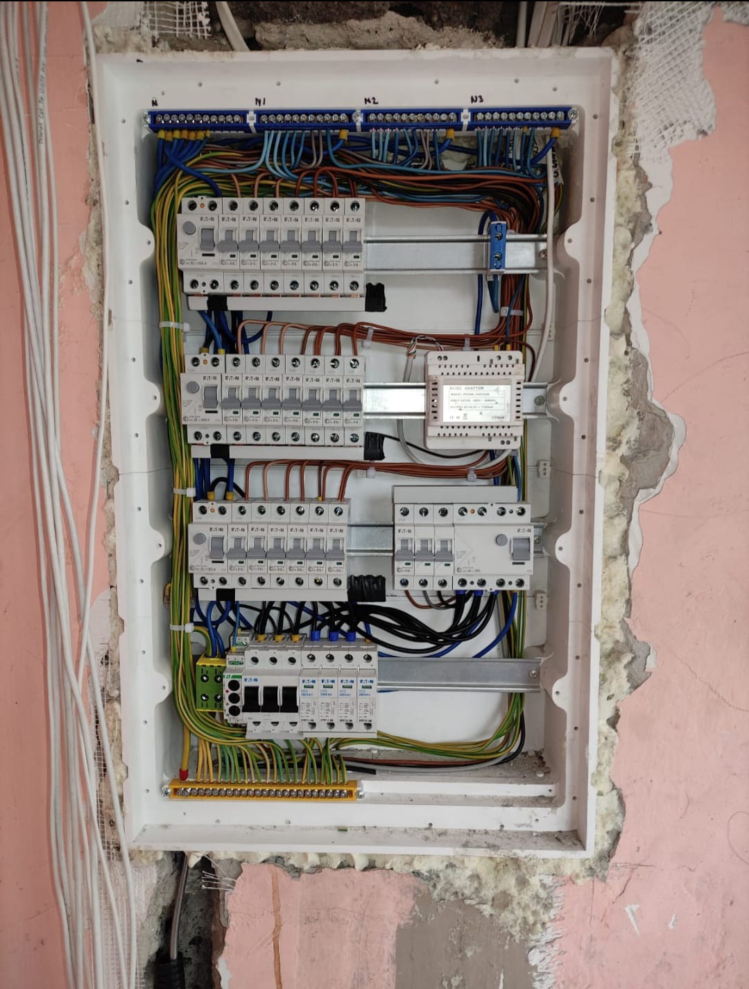 Otwarta skrzynka rozdzielcza z widocznymi bezpiecznikami, przewodami różnej grubości i kolorów, zamontowana w ścianie w trakcie remontu, z widocznymi śladami po kuciu i resztkami zaprawy.