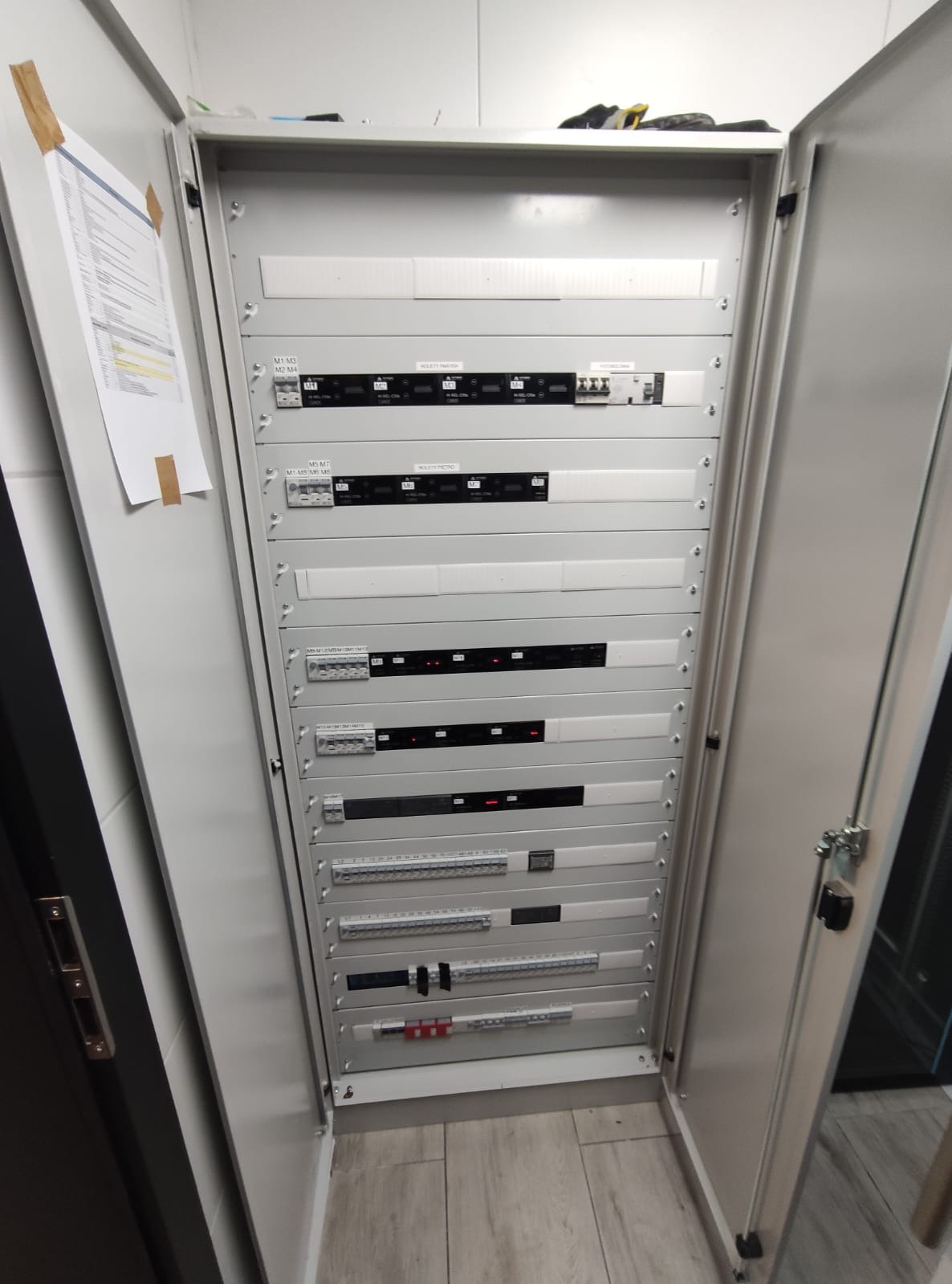 Otwarta szafa elektryczna z zamontowanymi modułami, widoczne zabezpieczenia, listwy zaciskowe i dokumentacja przyklejona do drzwi.