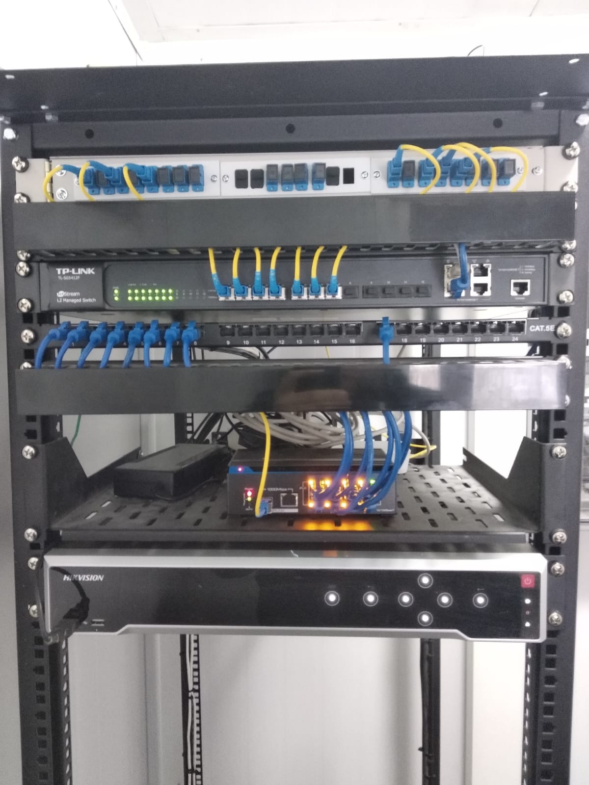 Zamontowana szafa rack z panelami krosowniczymi, switchem TP-Link i rejestratorem Hikvision, okablowanie strukturalne z niebieskimi i żółtymi kablami patchcord, diody LED sygnalizujące pracę urządzeń.