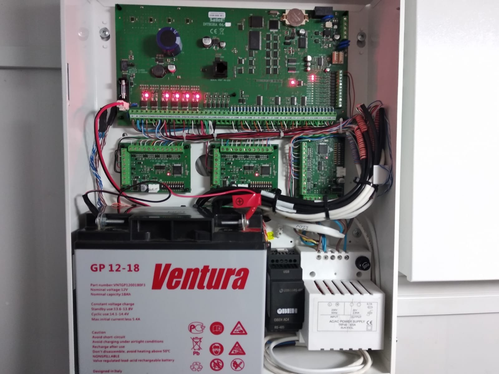 Otwarta obudowa centrali alarmowej z widoczną płytą główną, modułami rozszerzeń, akumulatorem podtrzymującym i zasilaczem, liczne przewody połączeniowe.