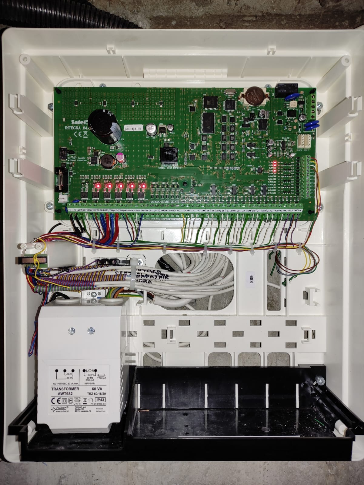 Otwarta obudowa centrali alarmowej Integra 64 z widoczną płytą główną, licznymi przewodami i transformatorem Pulsar AWT682, zamontowana w białej obudowie z czarnym spodem.
