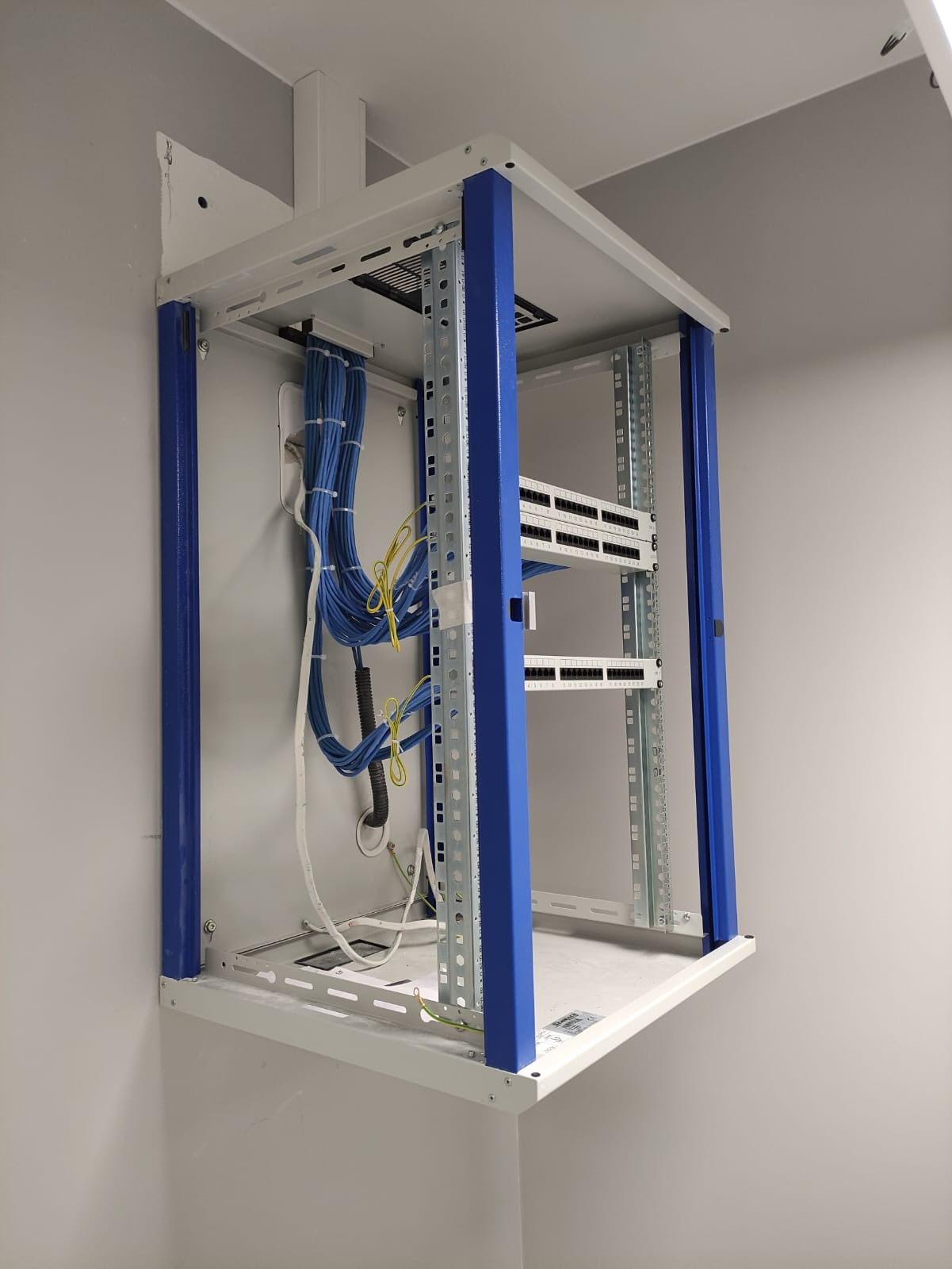 Półka rackowa z niebieskimi słupkami, zamontowana na ścianie, z widocznymi kablami sieciowymi i panelami krosowniczymi.
