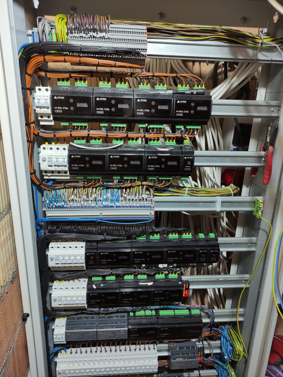 Szafa sterownicza z modułami przekaźnikowymi, bezpiecznikami i licznymi przewodami w różnych kolorach, uporządkowanymi w wiązki i podłączonymi do listew zaciskowych.