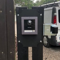 Balex electro - Zewnętrzny panel wideodomofonu Hikvision zamontowany na czarnym słupku bramy wjazdowej, widoczny mechanizm otwierania bramy i fragment białego samochodu dostawczego w tle.