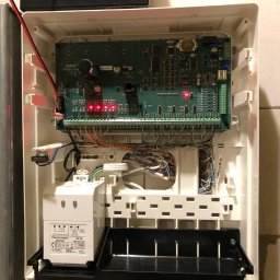 Balex electro - Otwarta skrzynka centrali alarmowej z widoczną płytą główną, licznymi przewodami i transformatorem na dole, zasilana akumulatorem Pb na górze