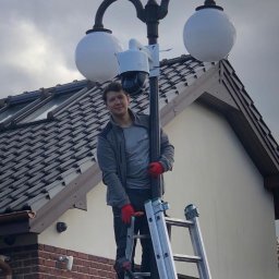 Balex electro - Mężczyzna na drabinie instaluje kamerę monitoringu na słupie oświetleniowym z dwoma kulistymi kloszami, widoczny budynek z dachówką.