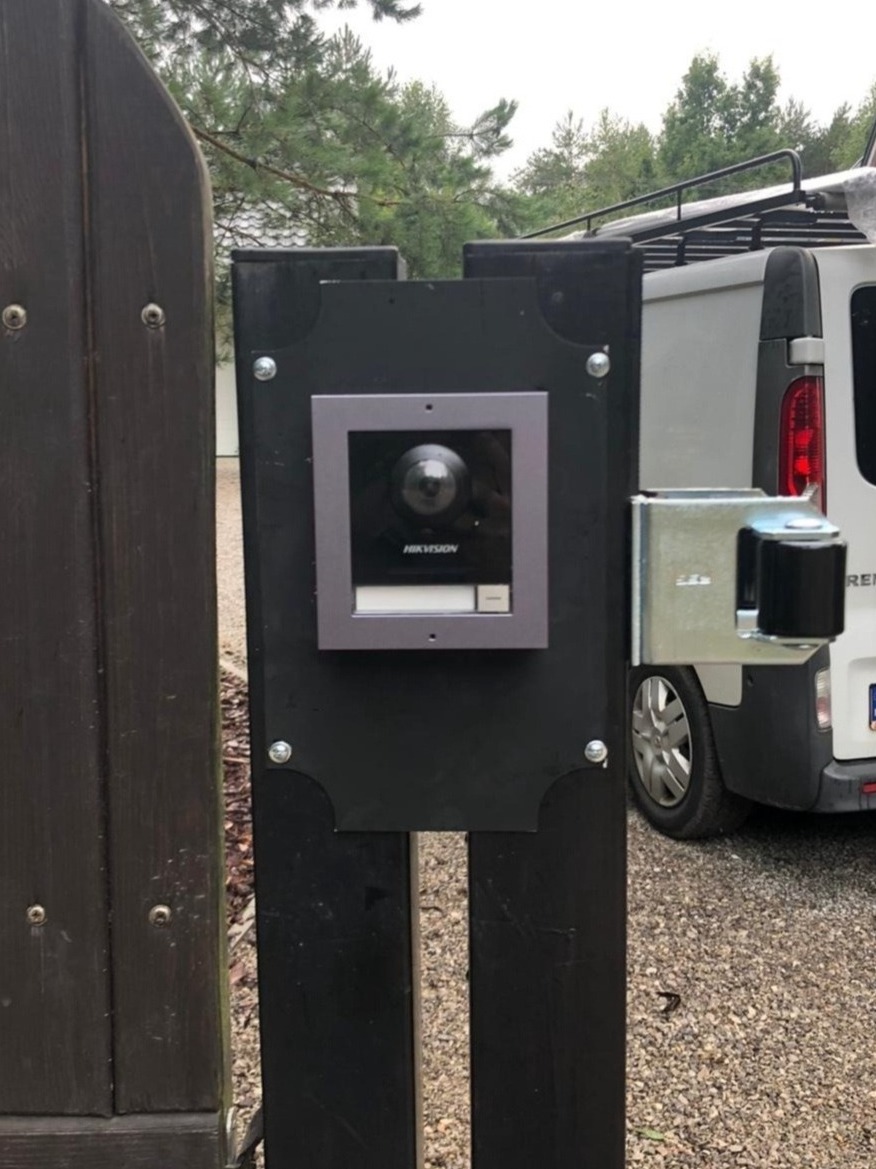 Zewnętrzny panel wideodomofonu Hikvision zamontowany na czarnym słupku bramy wjazdowej, widoczny mechanizm otwierania bramy i fragment białego samochodu dostawczego w tle.