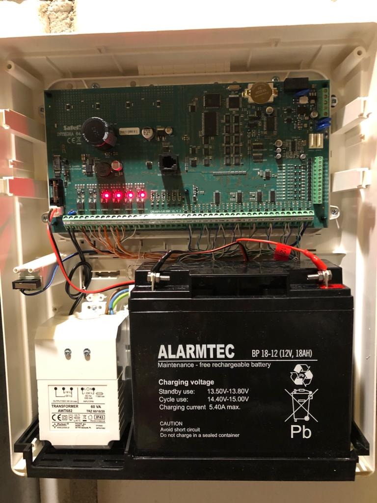 Wnętrze obudowy alarmowej z płytą główną, transformatorem i akumulatorem ALARMTEC BP 18-12, widoczne czerwone diody LED.