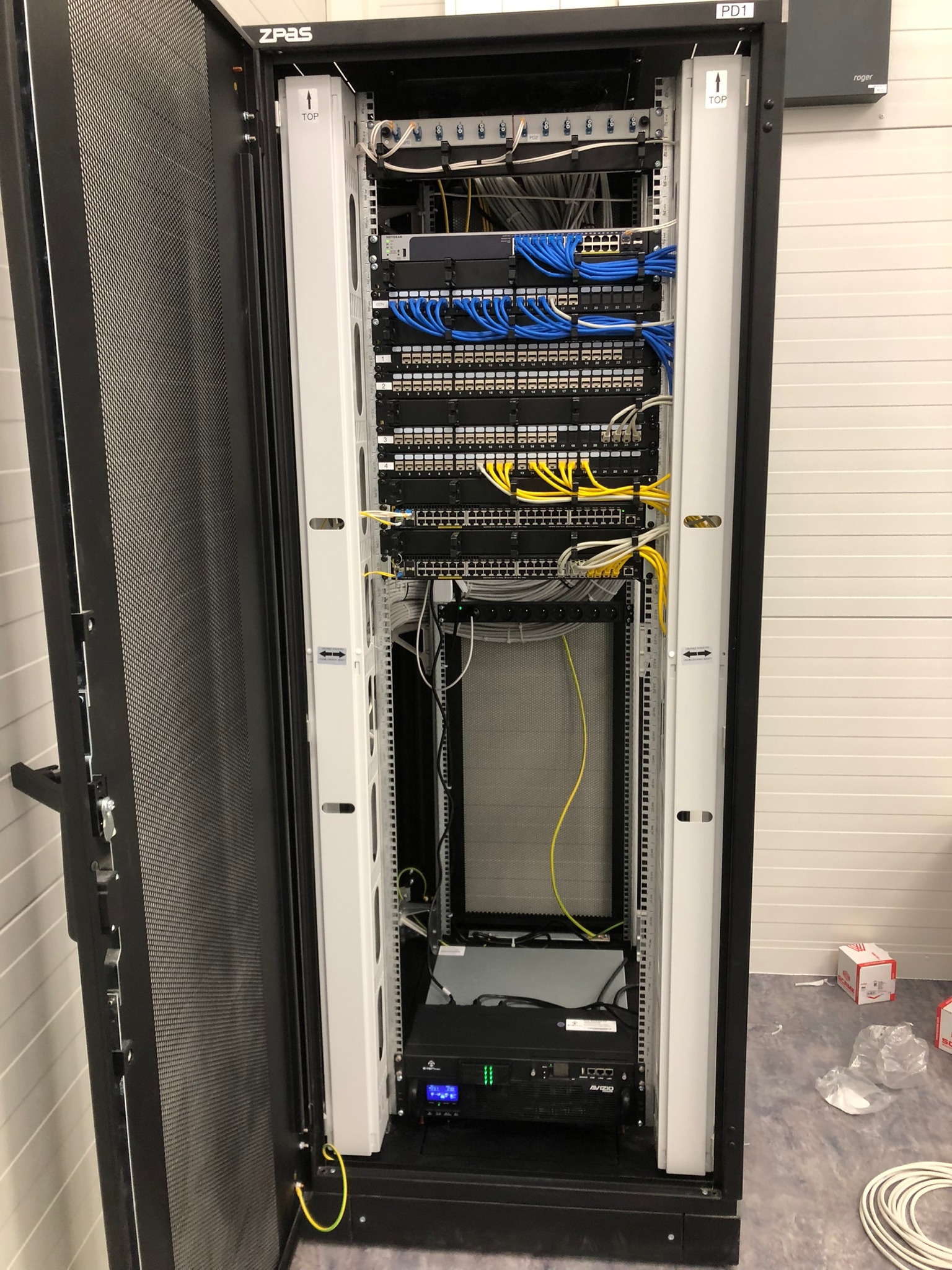 Otwarta szafa rack z zainstalowanym sprzętem sieciowym: switche, patch panele, okablowanie strukturalne w kolorach niebieskim i żółtym, widoczny kabel uziemiający.