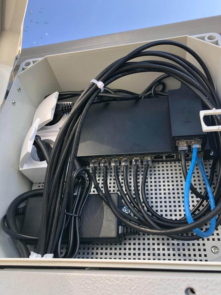 Wewnętrzny widok szafki z zainstalowanym switchem sieciowym, zasilaczami i kablami Ethernet, uporządkowanymi za pomocą opasek zaciskowych, umieszczonymi na perforowanej płycie montażowej w szarej...