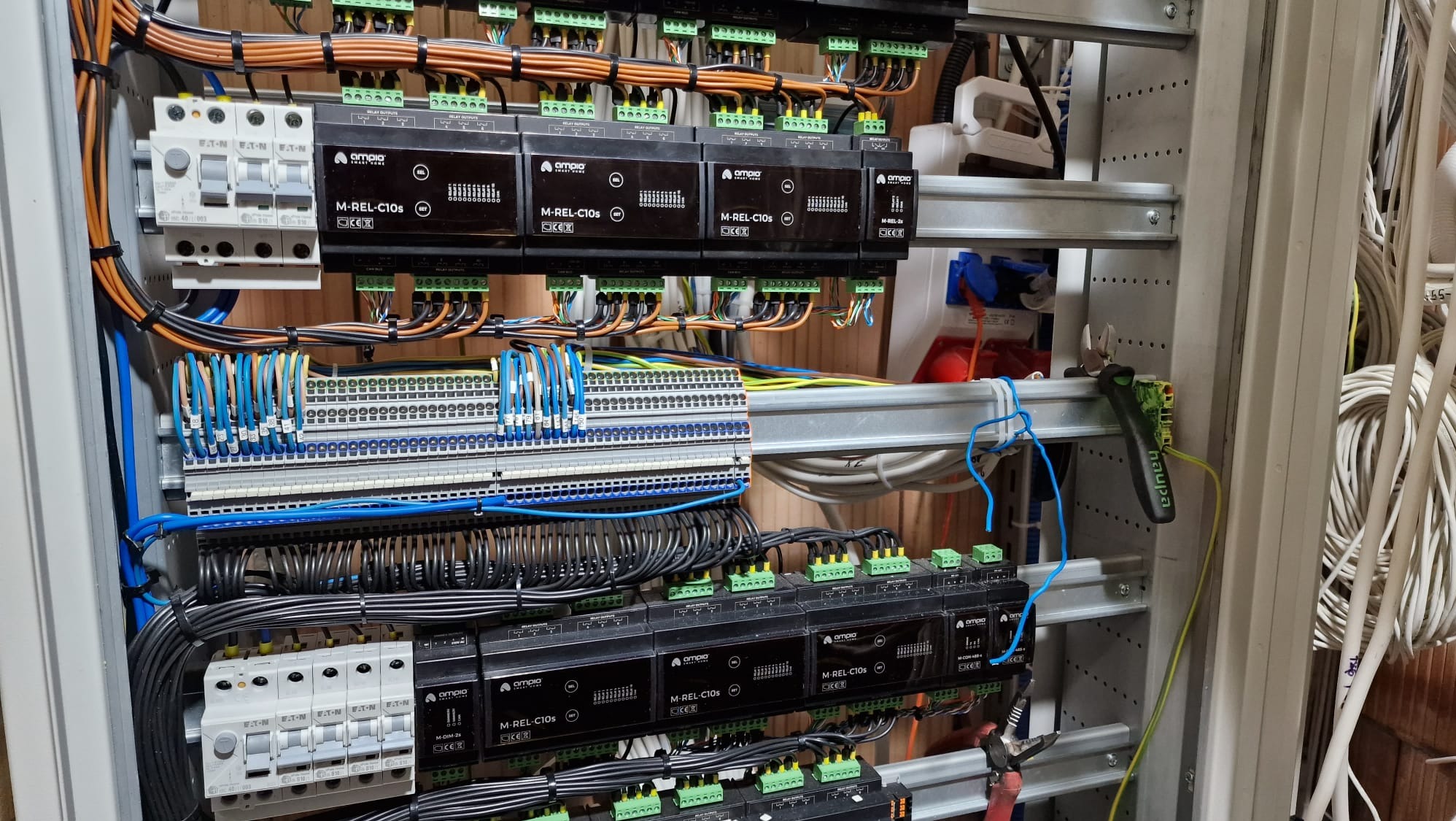 Rozdzielnica elektryczna z modułami sterującymi, licznymi przewodami w różnych kolorach, listwami zaciskowymi i bezpiecznikami, widoczne narzędzie do cięcia przewodów.