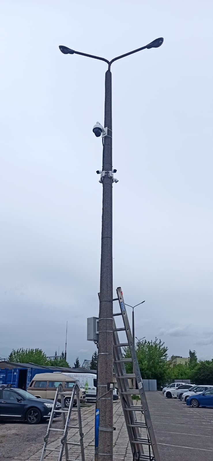 Słup oświetleniowy z zamontowanymi kamerami monitoringu, drabina oparta o słup, parking w tle.