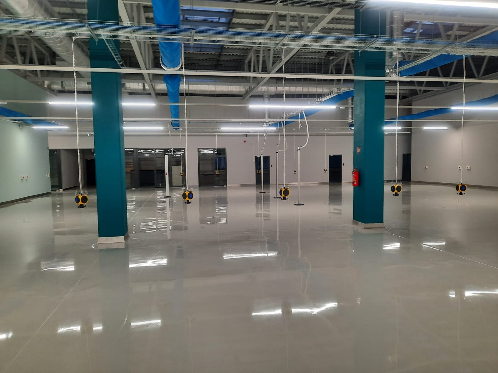 Wnętrze hali produkcyjnej z widoczną instalacją elektryczną pod sufitem, niebieskie rury wentylacyjne, oświetlenie LED i słupy pomalowane na niebiesko.