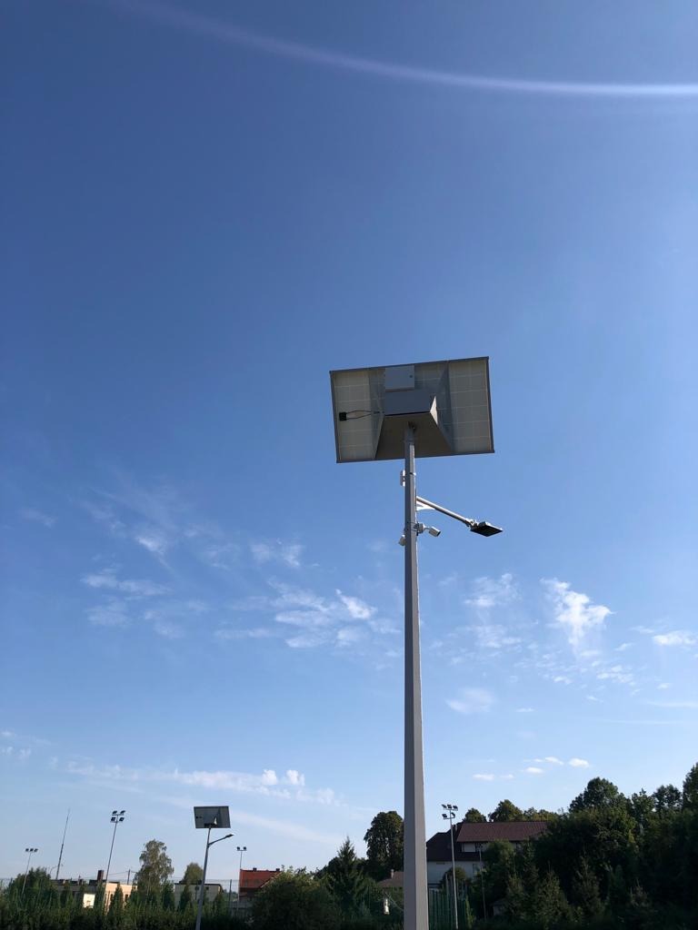Słup oświetleniowy z panelem solarnym i kamerami monitoringu na tle błękitnego nieba z delikatnymi chmurami, widoczne inne słupy oświetleniowe i zabudowania w oddali.