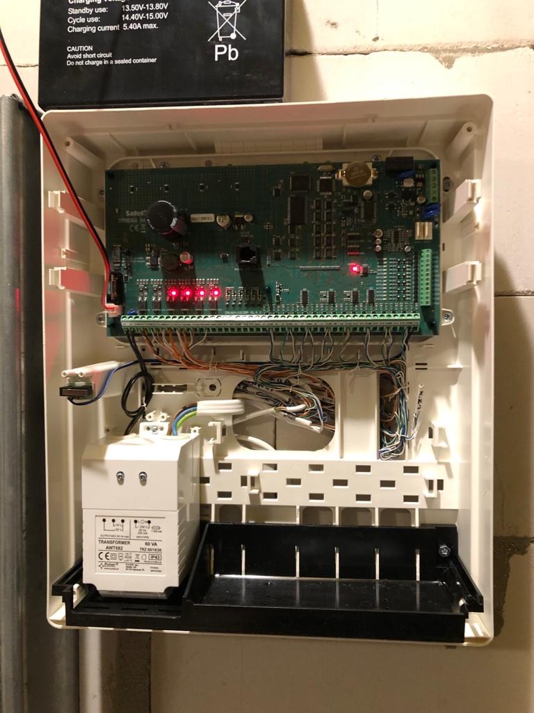 Otwarta skrzynka centrali alarmowej z widoczną płytą główną, licznymi przewodami i transformatorem na dole, zasilana akumulatorem Pb na górze