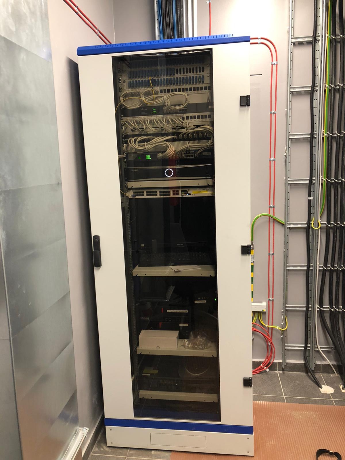 Szafa rack z zainstalowanym sprzętem sieciowym, widoczne patch panele, switche i okablowanie, kable prowadzone w korytkach na ścianie.