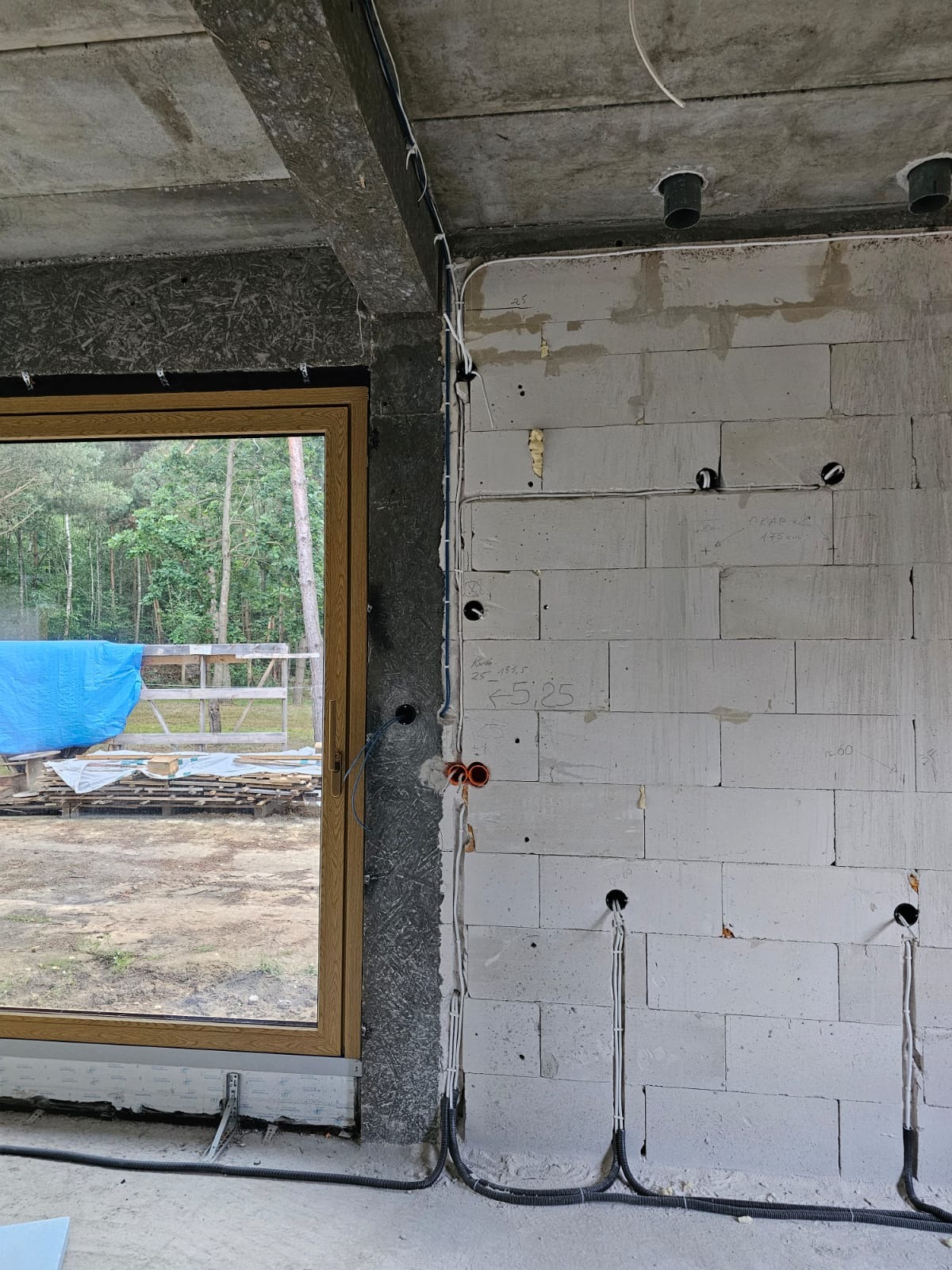 Surowy stan instalacji elektrycznej w nowym budynku: widoczne kable w peszlach prowadzone wzdłuż ściany z betonu komórkowego, otwory pod gniazdka, sufit betonowy z rurami instalacyjnymi, okno...