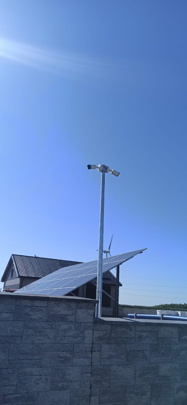 Zewnętrzny monitoring z dwoma kamerami na metalowym słupie, panele słoneczne na dachu budynku w tle oraz mała turbina wiatrowa, widok z dołu na błękitne niebo.
